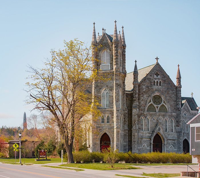 A grey stone church with trees beginning to show spring leaves in a downtown on a sunny day.