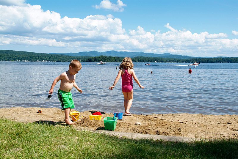 Two children play in the sand next to a lake in summertime.