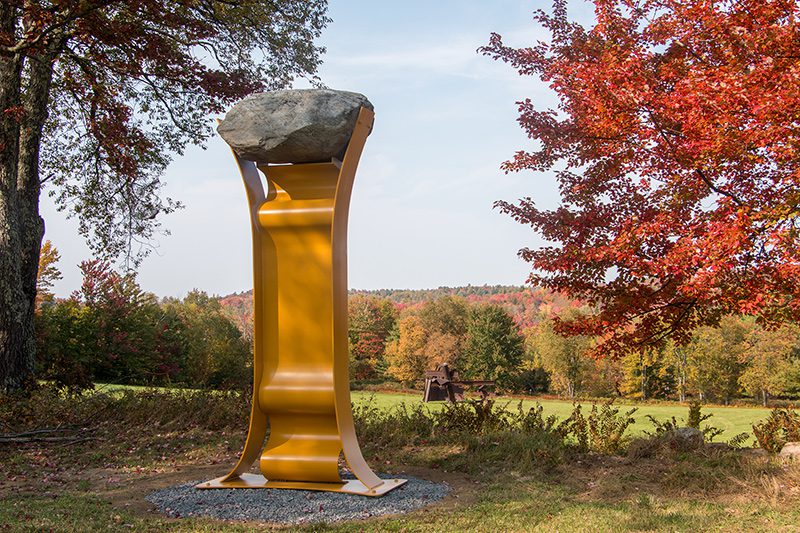 A golden art piece sits in an outdoor field with trees in fall foliage.