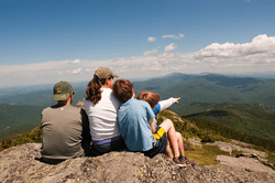 A family sits on the exposed rock of a mountain peak looking over the mountain valley on a sunny day.