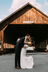 Two people kiss in front of a wooden covered bridge.