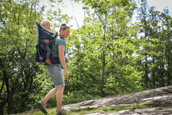 A person hikes through the forest in the summer with a baby on their back.