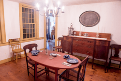 A historic living room with a fireplace, a table in the middle set for tea, and hardwood floors.