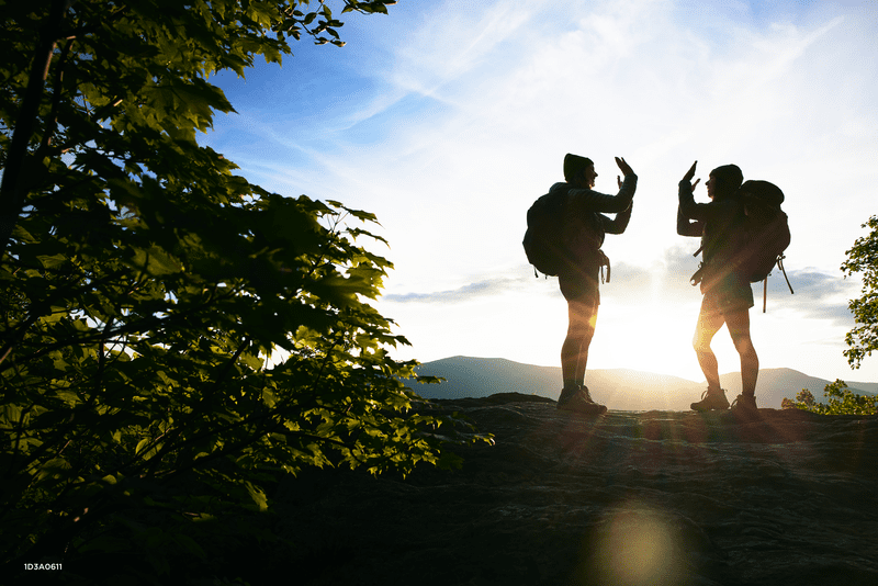 Two hikers wearing backpacks are silhouetted in front of the setting sun in the woods.