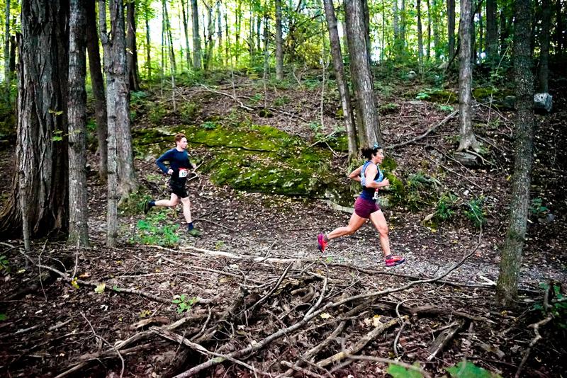 Two people running on a dirt hiking trail winding through a forest lined with blooming white trillium wildflowers.