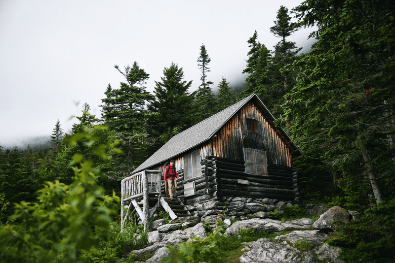 A person in a red jacket walks outside a remote cabin in the woods.
