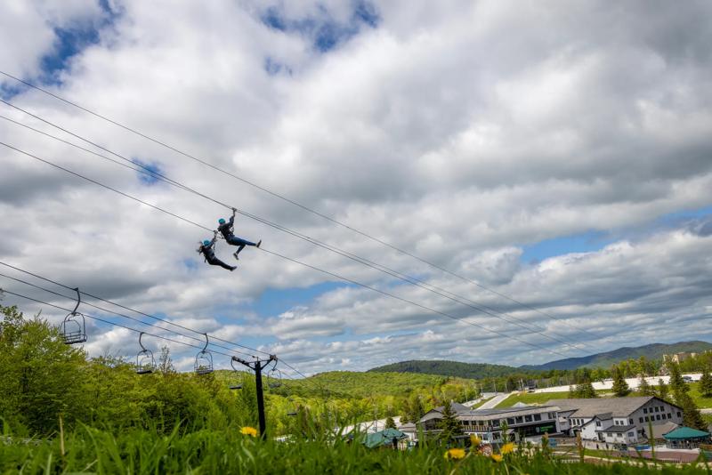 Two people ride ziplines high above a mountain resort with a ski lift, green hills, and a cloudy blue sky in the background.