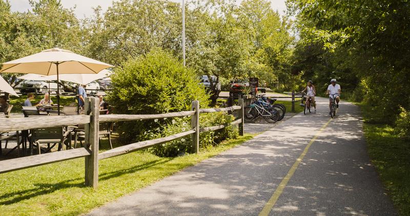 Two cyclists ride along a paved recreation path next to a wooden fence and an outdoor patio with umbrella seating.