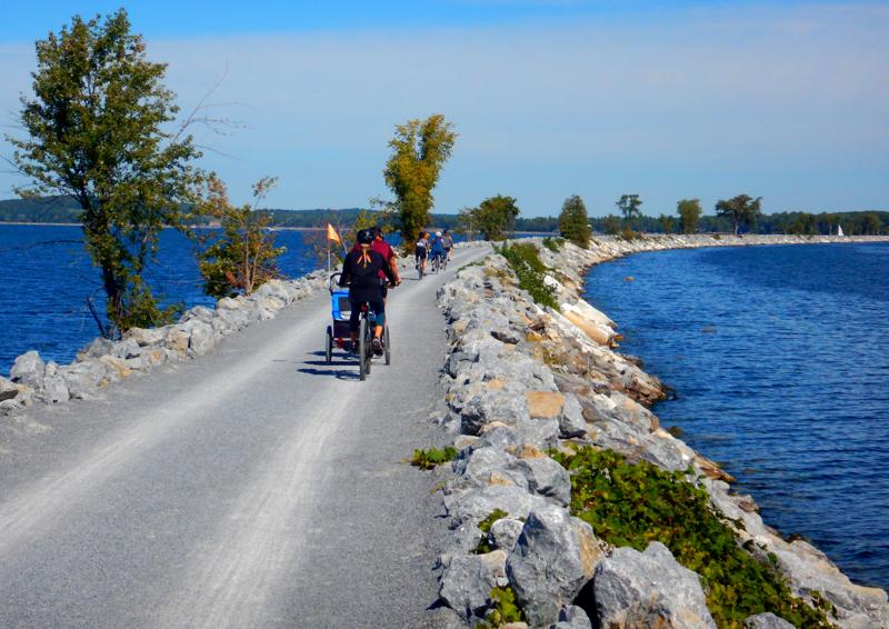 A family riding bicycles and pulling a child trailer along the paved Burlington Greenway path next to Lake Champlain.