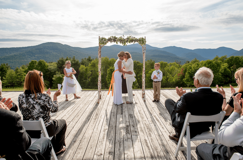 Two people kiss under an arch at an outdoor wedding with mountains visible behind.