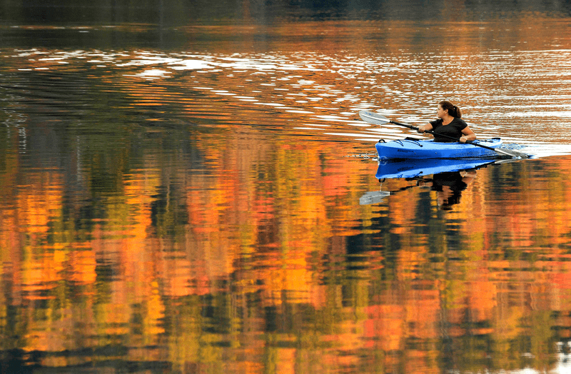 A person paddles a kayak across a calm body of water. The colors of fall foliage are reflected on the water.