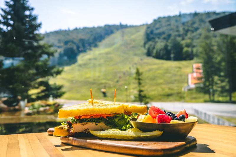 Outdoor dining table featuring a sandwich, pickle, and bowl of fruit with scenic views of summer ski slopes and mountain bike trails in the background.