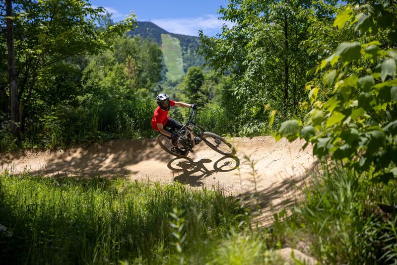 A mountain biker wearing a helmet and red shirt navigating a curved dirt trail, with a sunny ski resort slope framed by trees in the background.