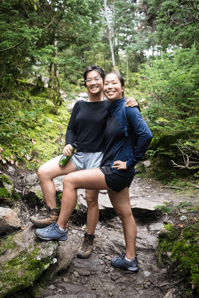Two people pose for the camera on a narrow, dirt hiking path in the woods.