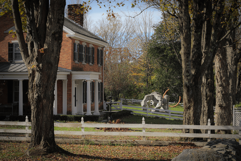 A side view of a historic red brick house sitting next to trees. There is an abstract sculpture in the front yard.