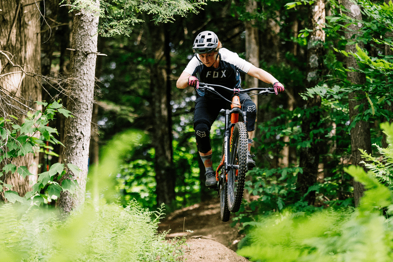 A person rides a mountain bike toward the camera out of a forest in the summer.