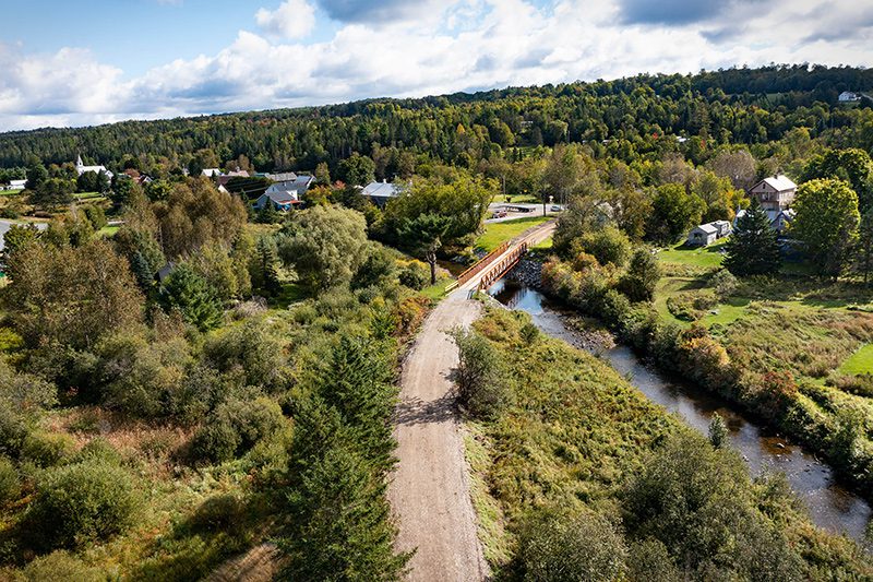 Seen from above, a gravel path with a bridge stretching across a river in summer.
