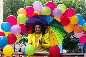 A person wearing a bright yellow outfit and holding a rainbow patterned umbrella stands underneath a colorful balloon arch.