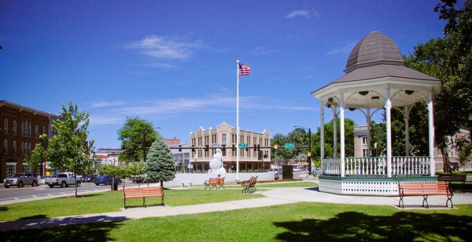 A park with a gazebo, flag, stone statue, and buildings in the background is seen on a warm, sunny day.