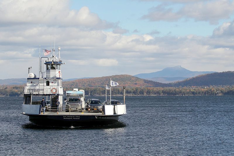 Cars sit on a small ferry as it moves across a lake. A mountain ridge sits in the distant background.