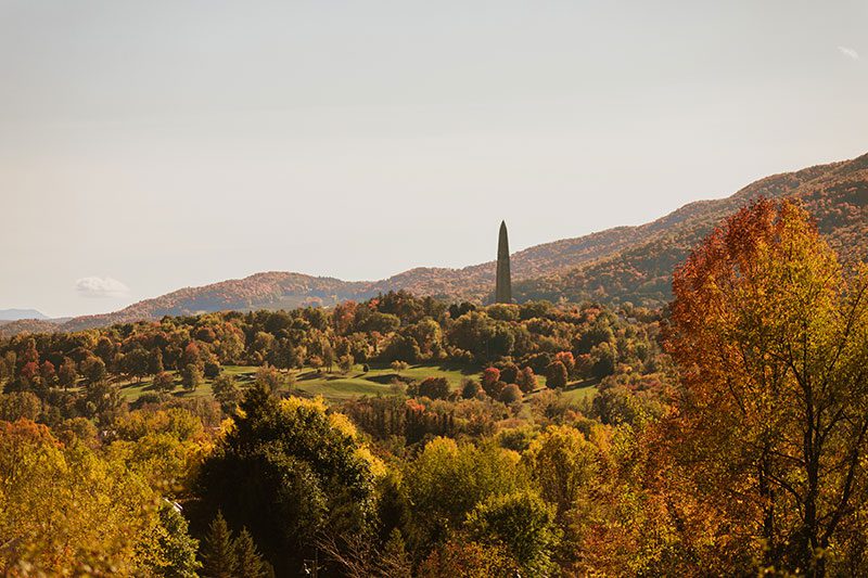 A tall monument is seen from afar, rising above trees in the autumn.