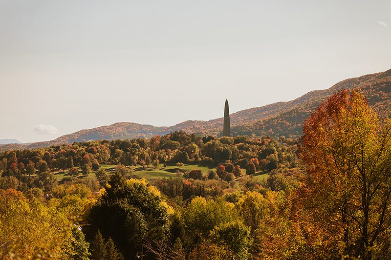Seen from above, the view of colorful fall leaves on hills and mountains. A tall monument sits in the distance.