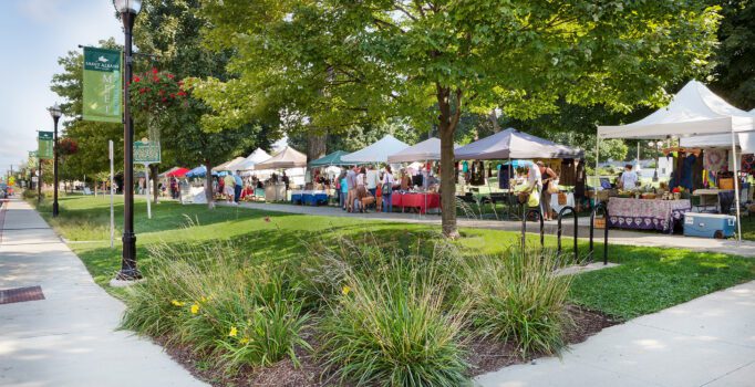 A row of vendor tents line a street at a farmers market on a warm, sunny day.