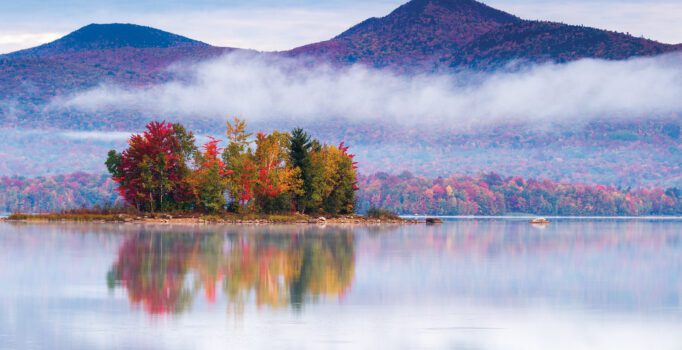 Low-hanging clouds form at the base of two mountains and just above a lake with a small tree-covered island. The sun reflects the foliage on the trees in the water.