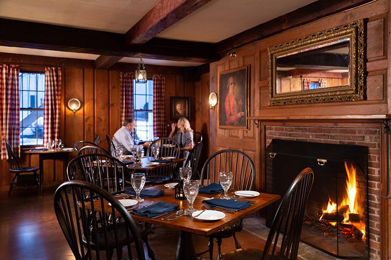 Two people dine in a wood-paneled tavern with warm wooden tables and chairs and a fire in a fireplace.