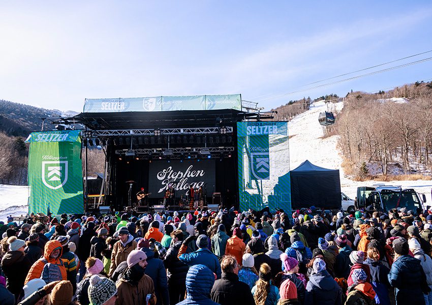A crowd of people watch live music on a stage at the base of a snow-covered ski trail.