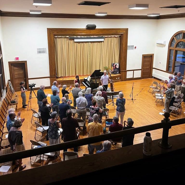 A crowd of people watch a concert in an indoor hall with wooden floors.