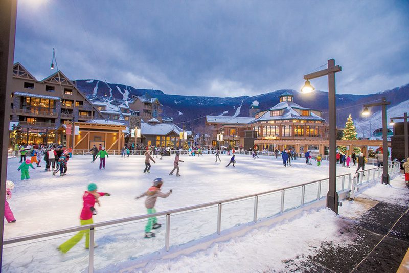 People ice skate at an outdoor rink in the middle of resort buildings lit with a warm glow.