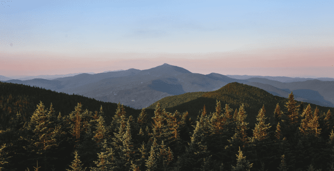 A view from above of a forest and rows of mountain peaks. The sky at the horizon is orange.