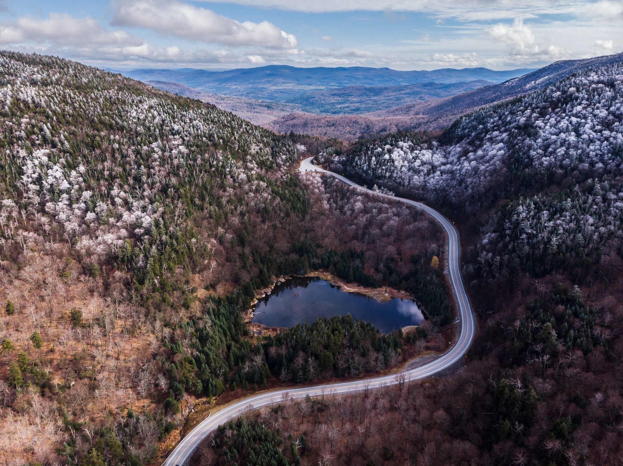 Seen from above, a winding road traverses through a mountain pass with a lake and snow-covered trees.