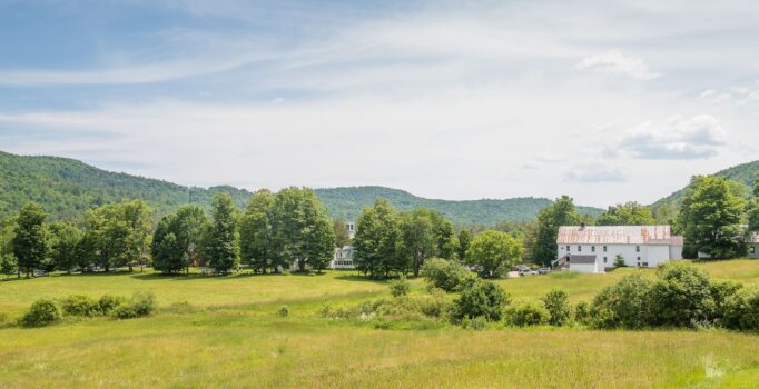 A white building is situated across a green field and surrounded by green trees on a warm and sunny day.