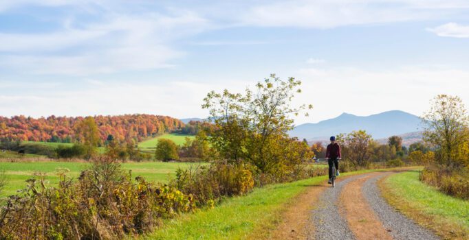 A person bikes on a gravel road lined with green grass. Fall foliage and a mountain are in the background.