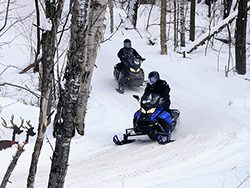 Two snowmobiles travel along a snowy trail in the woods.