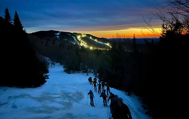 A group of people head uphill at a Vermont ski resort at night with bright lights illuminating the trail.