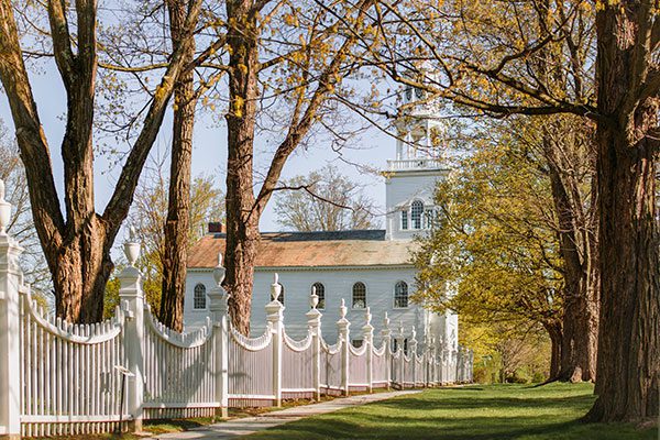 A white fence leading to a white church is surrounded by green trees on a sunny day.