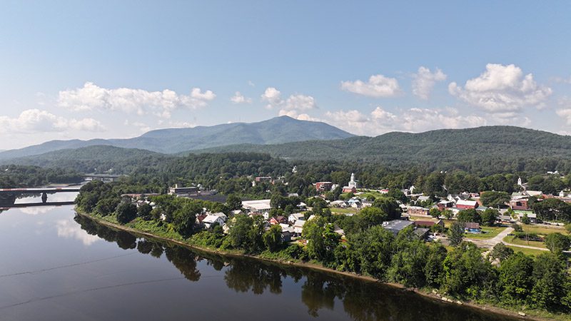 As seen from above, a rural town is surrounded by green trees, mountains, and a river on a warm sunny day.