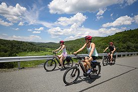 Three people ride bikes on a paved road with lush green mountains behind them.
