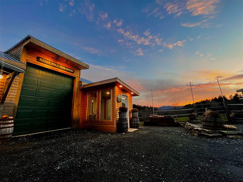 A wooden building with a sign that reads Bent Hill Brewery is lit up against the setting sun.