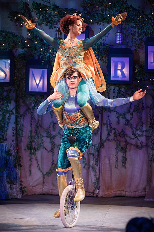 Two circus performers dressed in bright shiny colors on a unicycle, one on the shoulders of the other.