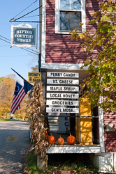 The front exterior of a country store on a fall day. Wood signs advertise products and pumpkins sit on the porch.