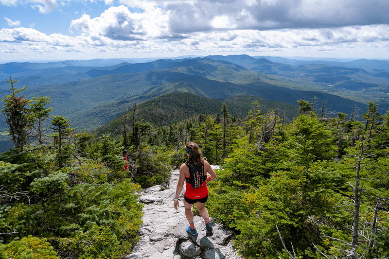 A hiker descends a mountain trail on a sunny day. Ahead of them is an expansive view of mountains in the distance.