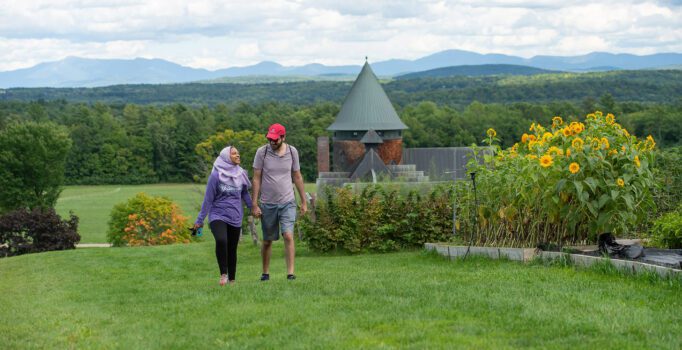 Two people walk across a grassy field toward the camera while a historic building is visible in the background.