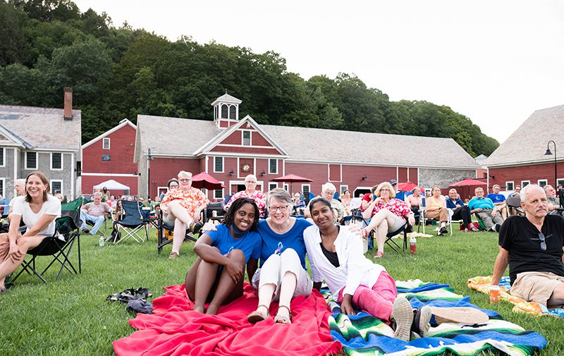 View of a group of people sitting on a green lawn with blankets, chairs, and a barn in the background.