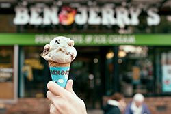 A scoop of ice cream on a cone held by a hand outside. In the background, a sign reads Ben & Jerry’s on a factory building.