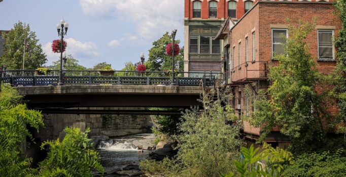 A bridge crosses a river in a town's downtown. Historic brick buildings line the bridge.