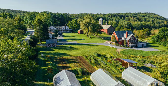 Seen from above, a farm with three greenhouses, a barn, and two houses. The sun is shining and green grass covers the farm.
